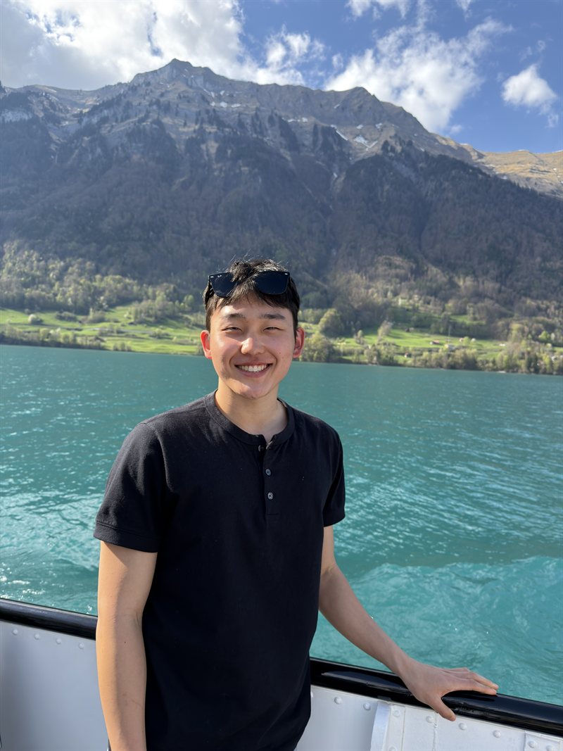 caleb kim smiling on a boat railing, a turquoise alpine lake and mountains behind him